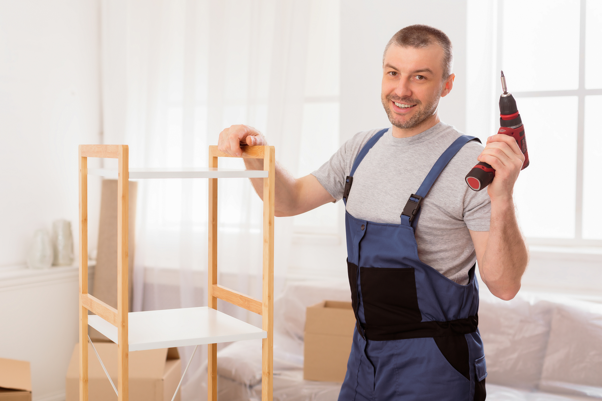 Handwerker im Overall hält Akkuschrauber und steht neben teils aufgebautem Holzregal in hellem Raum.